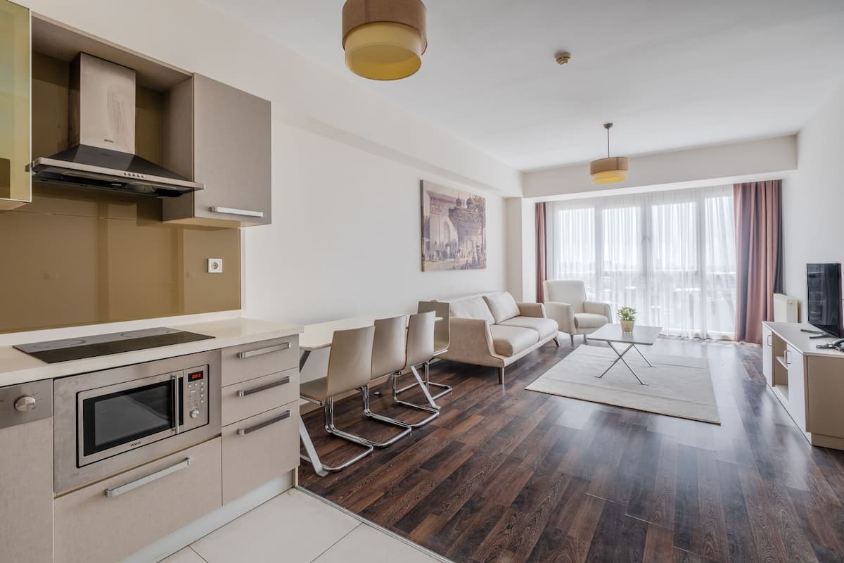 A modern living room with a sofa, coffee table, and TV. There is a kitchen area with a stove, oven, and cabinets.