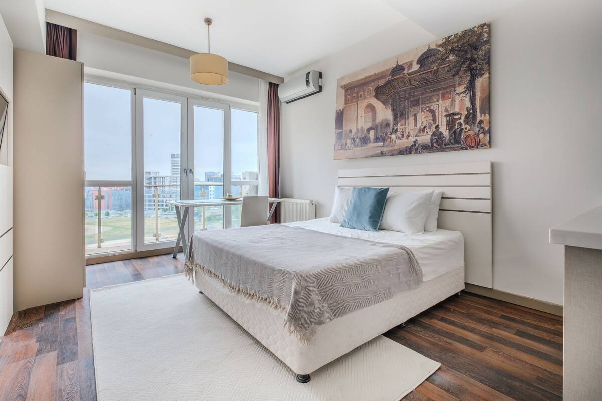 A modern bedroom with a large bed, a desk, and a view of a cityscape through large windows.