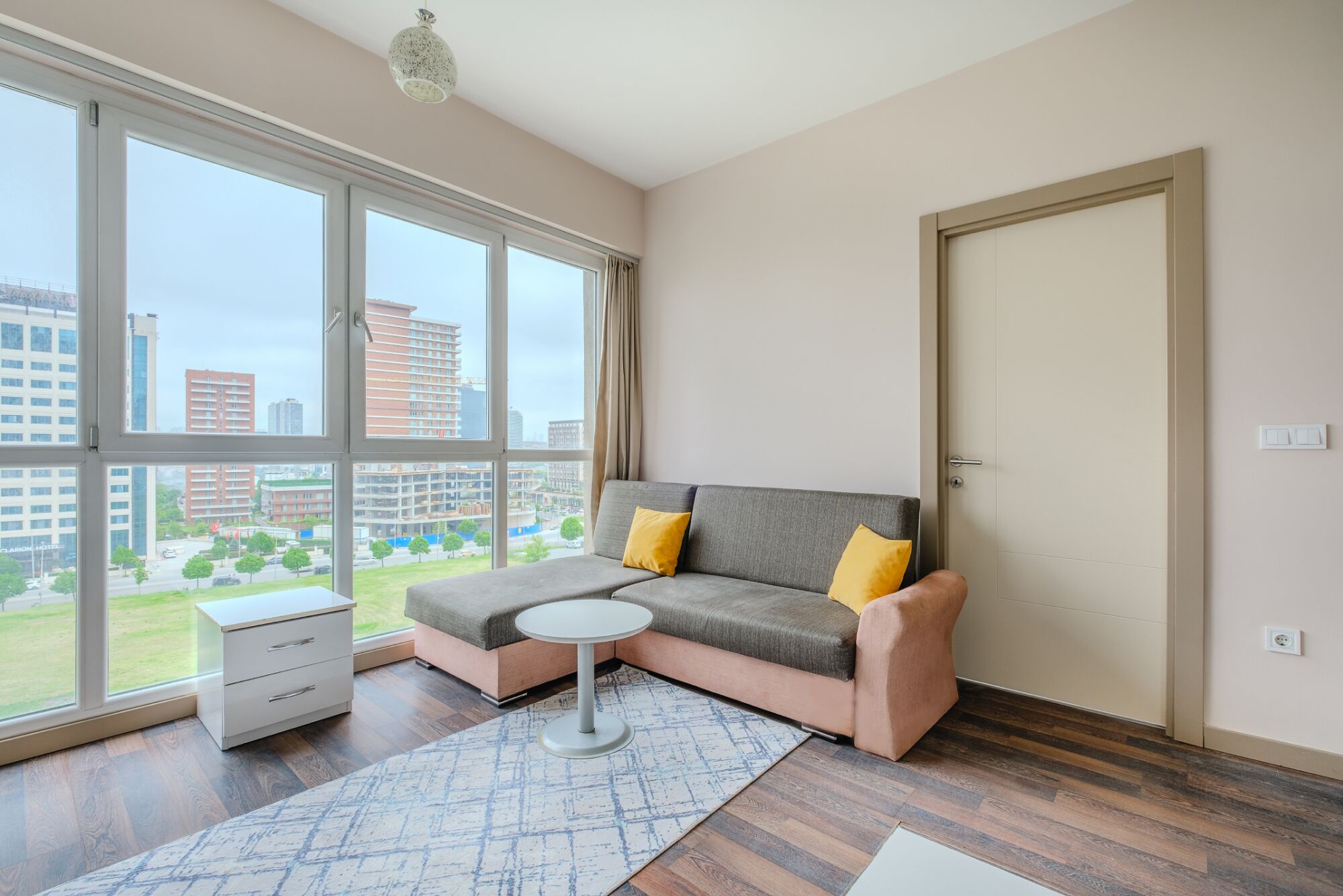 A modern living room with a grey sofa, two yellow cushions, a small round table, and a white side table. Large windows offer a view of buildings and a green area outside.