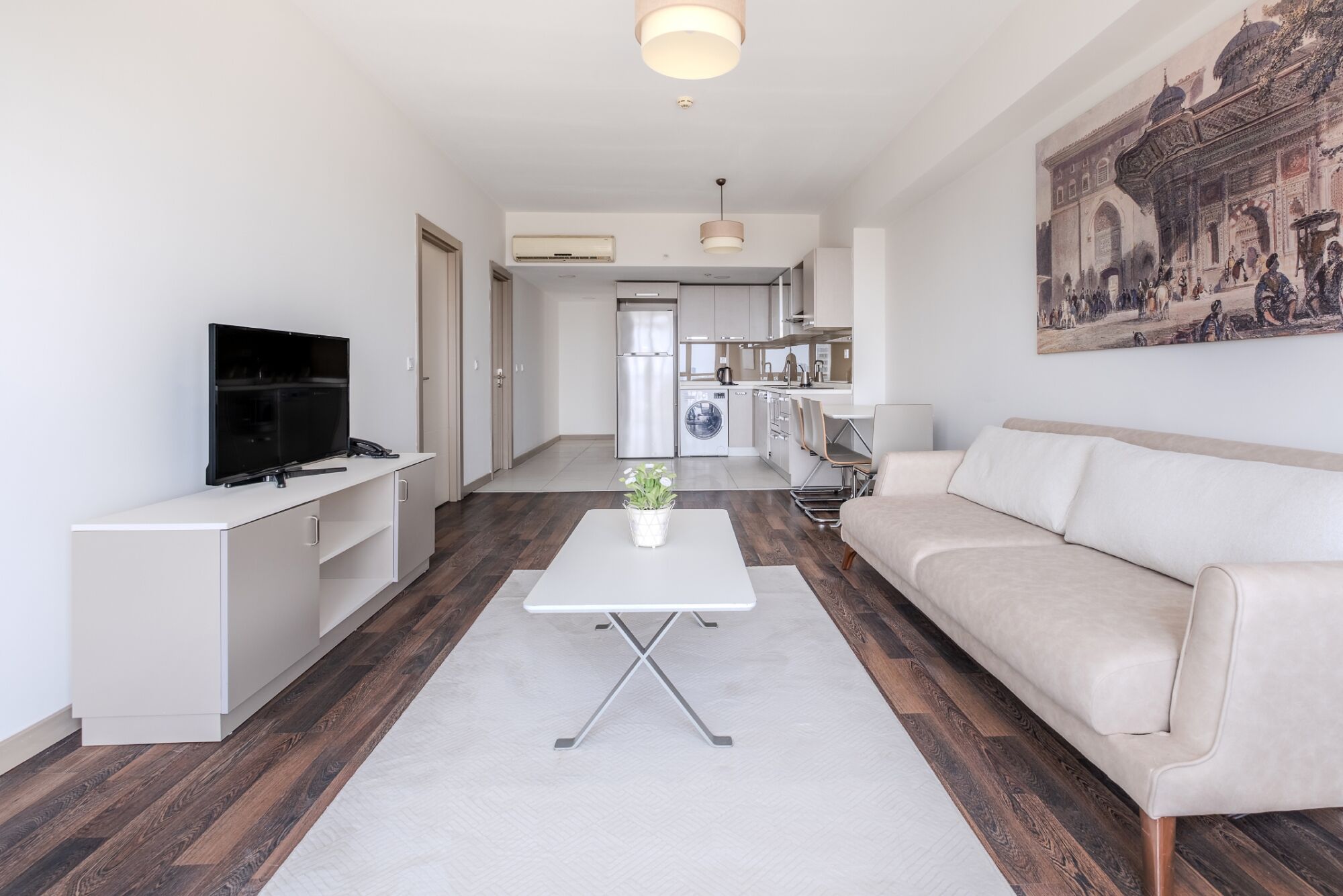A modern living room with a sofa, a coffee table, and a TV. There is a kitchen area with a washing machine and a dining table.