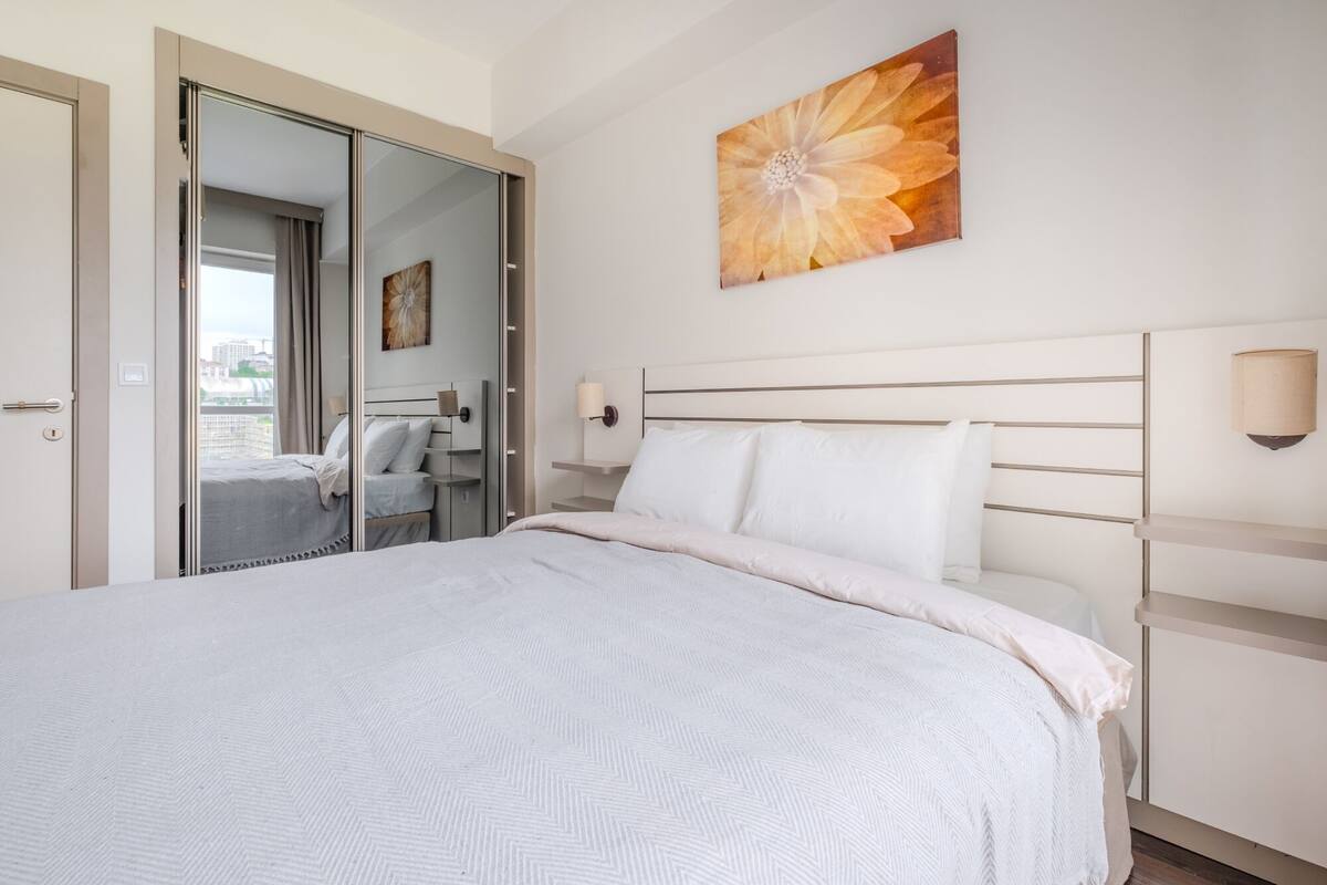 A modern bedroom with a large bed, a mirror, a wall-mounted flower painting, and a view of the city through a sliding door.