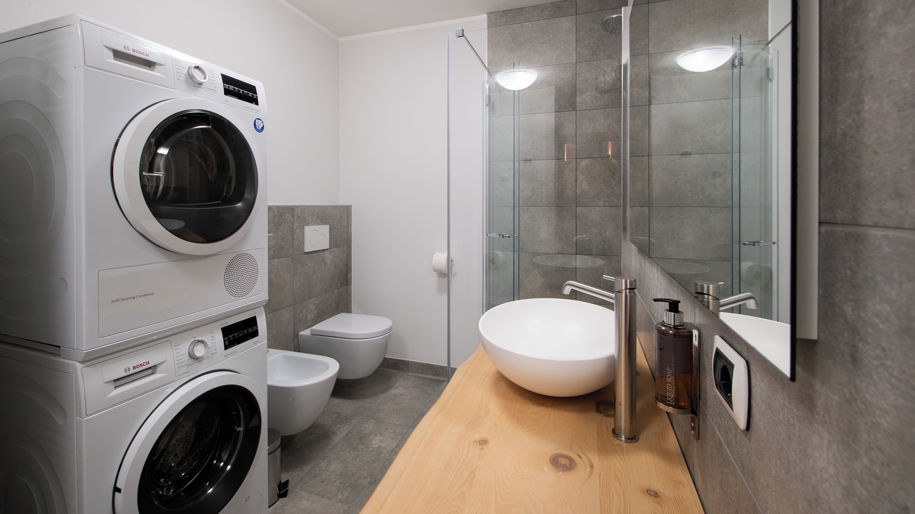 A modern bathroom with a Bosch washing machine, a toilet, a glass shower enclosure, and a wooden vanity with a sink.