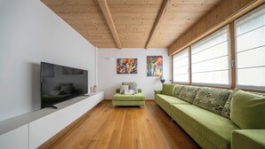 Een moderne woonkamer met een houten plafond, een flatscreen televisie, groene banken en grote ramen.