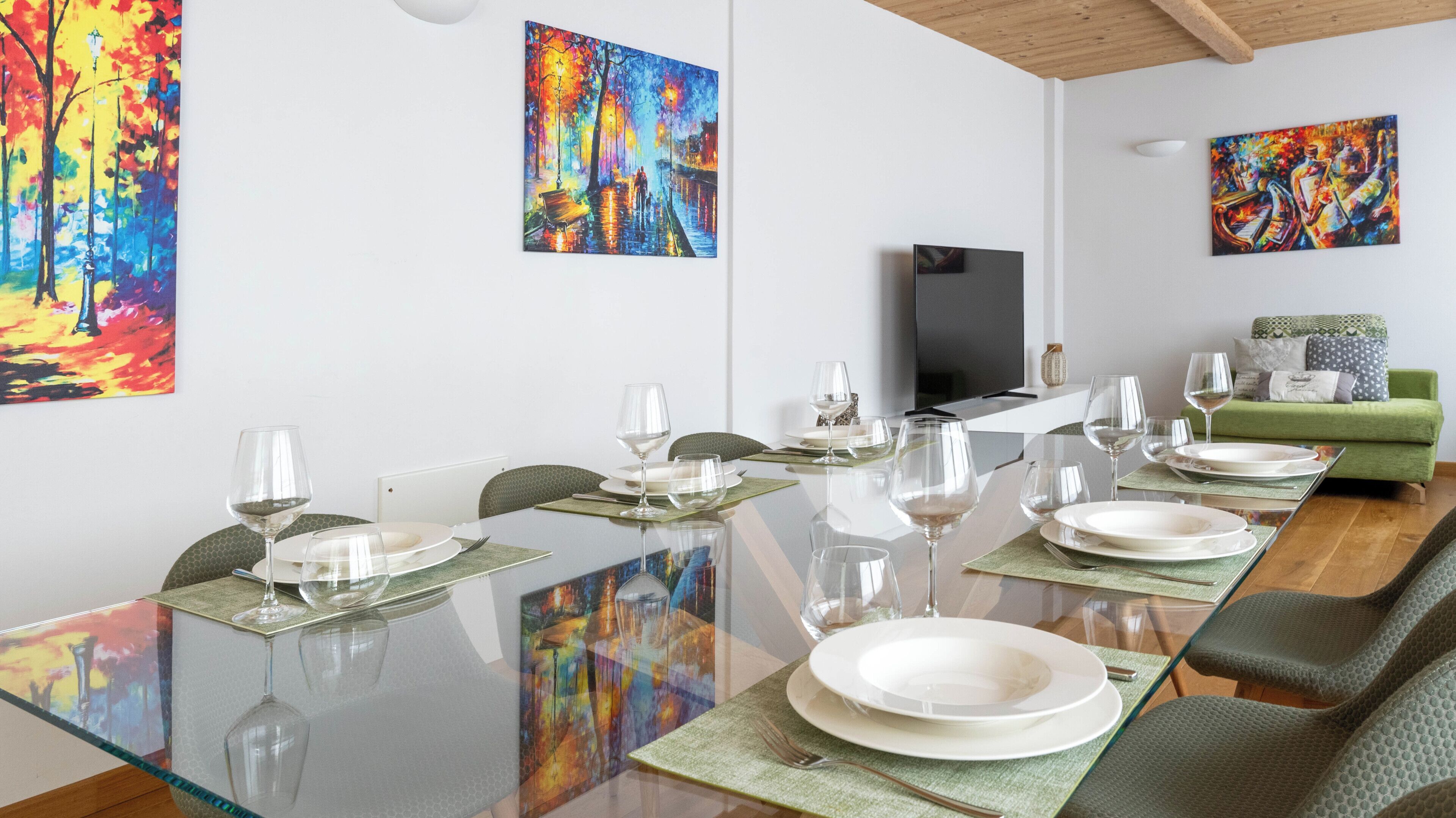 A dining area with a long table set for six, a television, and a green sofa in the background.