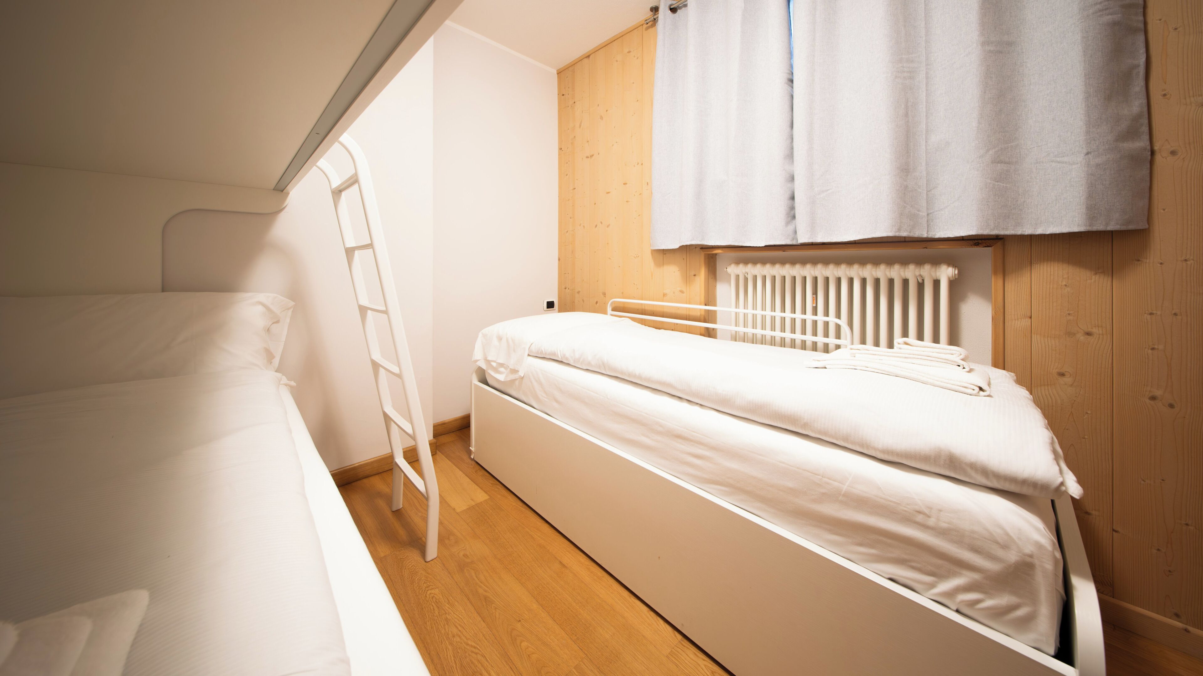Pequeña habitación con cama individual, suelo de madera, cama y escalera blancos, y un radiador.