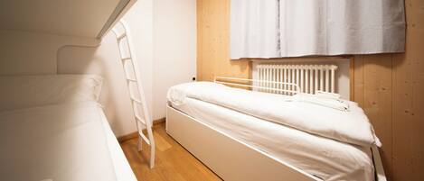 A small, single-bed room with a wooden floor, a white bed, a white ladder, and a radiator.