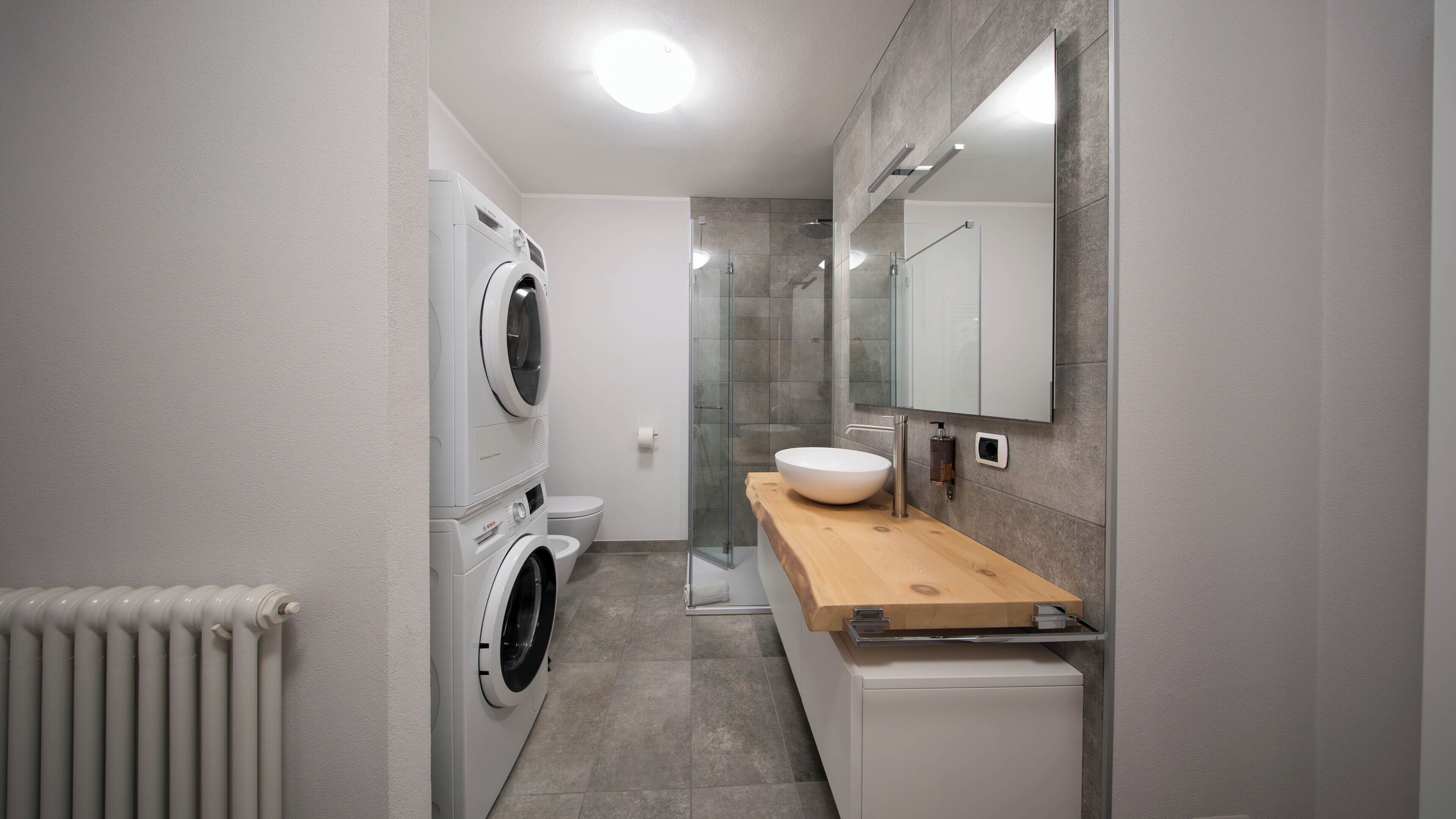 A compact bathroom with a washing machine, a toilet, a sink with a wooden countertop, and a glass shower enclosure.
