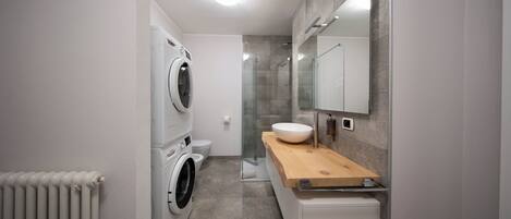 A compact bathroom with a washing machine, a toilet, a sink with a wooden countertop, and a glass shower enclosure.