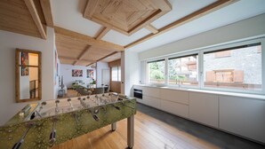 Een moderne keuken met een tafelvoetbaltafel, houten balken, grote ramen en uitzicht op een stenen gebouw.