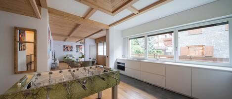 A modern kitchen with a foosball table, wooden beams, large windows, and a view of a stone building.