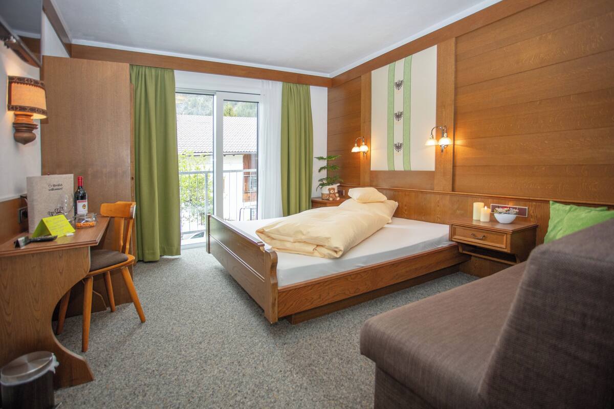 Ein Hotelzimmer mit einem hölzernen Bett, einem Schreibtisch mit Stuhl, einem Nachttisch mit Lampe und Blick ins Freie durch eine Schiebetür.