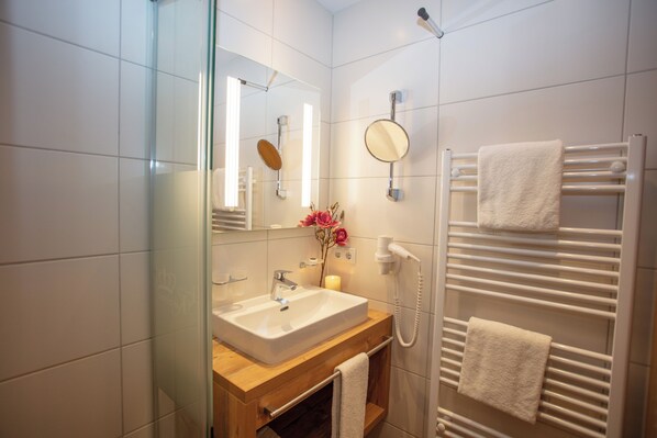 Un bagno moderno con box doccia in vetro, un lavandino bianco con piano in legno, uno specchio, un porta asciugamani e un soffione doccia manuale.
