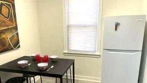A small kitchen with a white refrigerator, a dining table with four chairs, and a window with blinds. - The Kennywood Suite - The Blue Racer Car (Duquesne)
