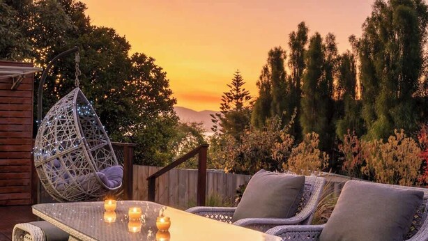A wicker outdoor seating area with a glass table and candles, surrounded by trees and a wooden fence.