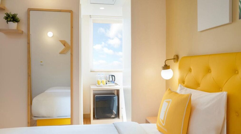 A hotel room with a bed, a yellow headboard, a bedside lamp, a window, and a small kitchenette area.