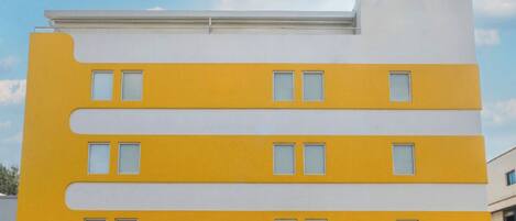 Un edificio moderno con fachada amarilla y blanca, con múltiples ventanas y un llamativo letrero 'bloom' en la parte superior.