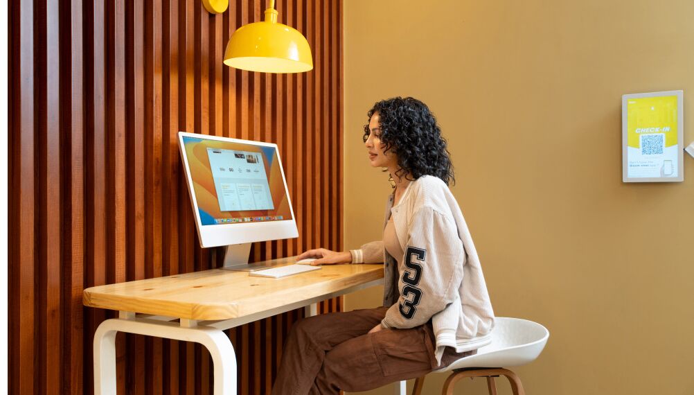 Una persona sentada en un escritorio de madera con una computadora, una lámpara colgante amarilla y un código QR en la pared.