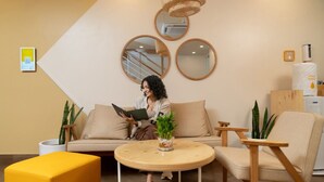 Egy modern szállodai előcsarnok, melyben egy kanapé, egy kerek, fából készült asztal és két szék található. A falon virágos növények és egy nagy tükör díszel.