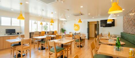 A modern dining area with wooden tables and chairs, yellow pendant lighting, and a brick wall.