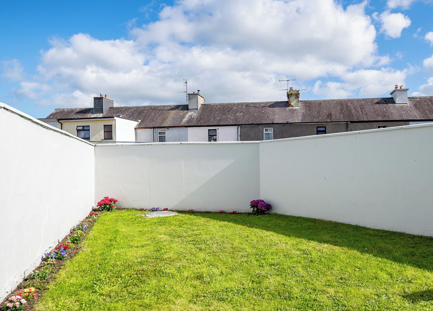 A small, enclosed garden area with a grassy lawn and flower beds, surrounded by a white wall.