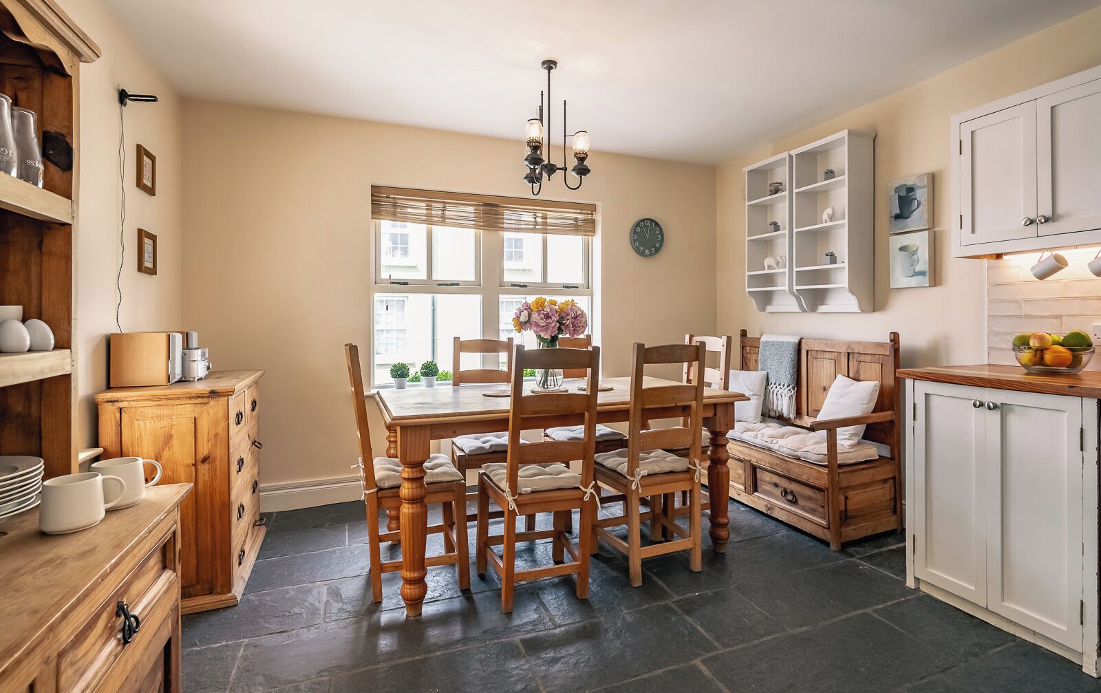 Una cocina acogedora con una mesa de madera, sillas y una ventana con persianas. Hay armarios, un reloj y una estantería con objetos.