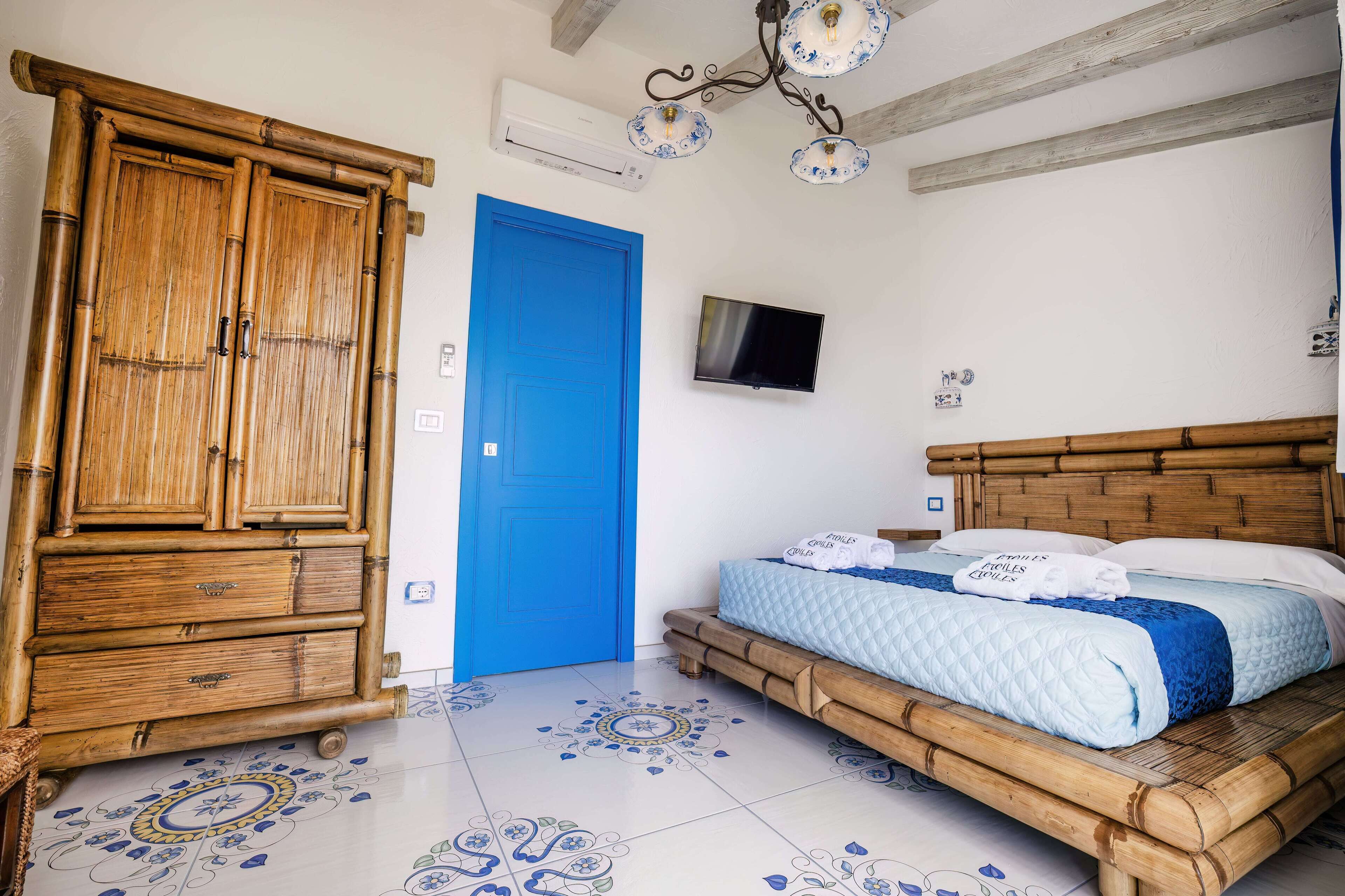A room with a blue door, a bed with a blue and white quilt, a wooden wardrobe, and a television on the wall.