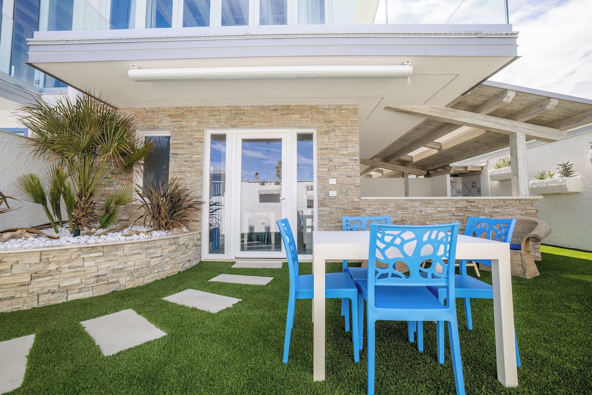 A patio with a white table and blue chairs, surrounded by a stone wall and palm plants.