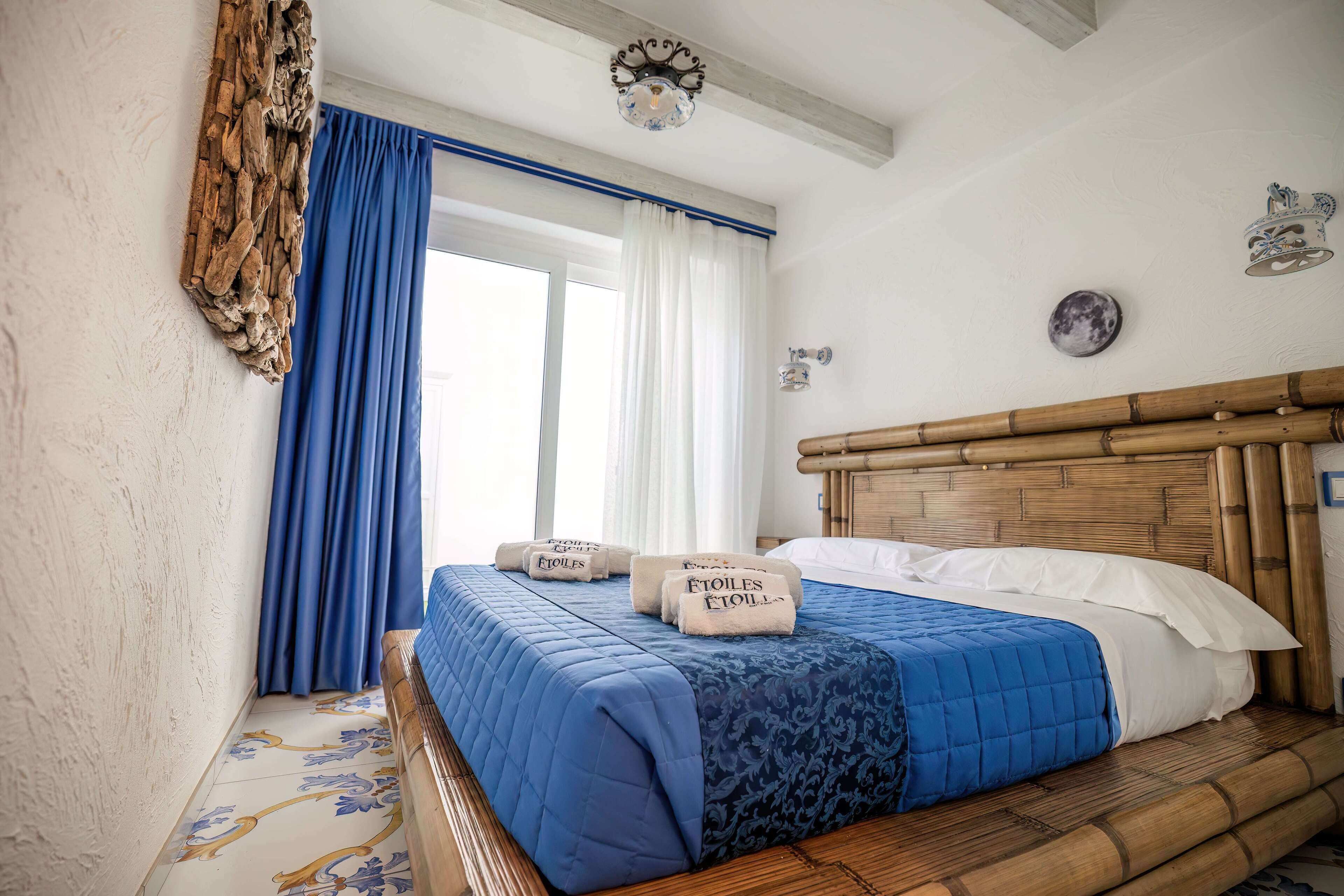 A neatly made bed with blue bedding and two towels, a wooden headboard, and a large window with blue curtains.