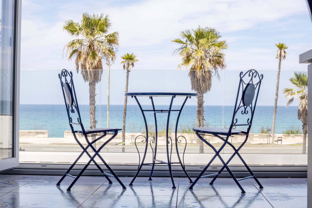 A balcony with two chairs and a table, overlooking the sea and palm trees.