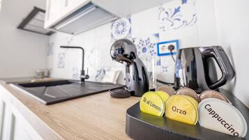 En køkkenbordplade med kaffemaskine, elkedel og stempelkande. Der er kaffekapsler med påskrifterne "Tê al Limone", "Orzo" og "Cappuccino".