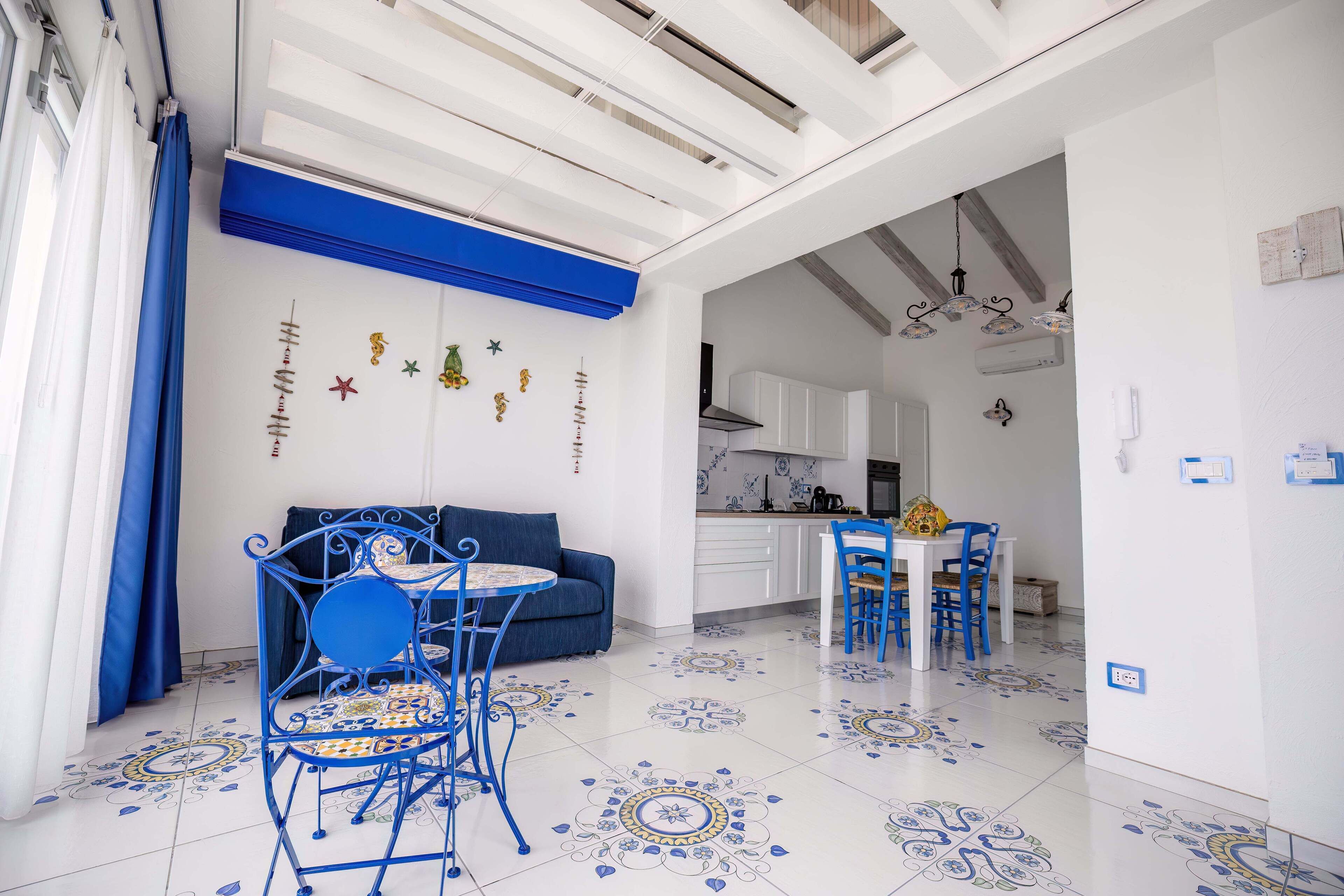 Una sala de estar moderna con un sofá azul, una mesa redonda y sillas azules a juego. La habitación presenta paredes blancas, detalles en azul y baldosas de suelo estampadas.