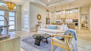 A spacious living area with a sofa, coffee table, and dining table. There is a kitchen in the background with modern appliances. - Stylish New Build with Pool, Spa, and Design (North Port)