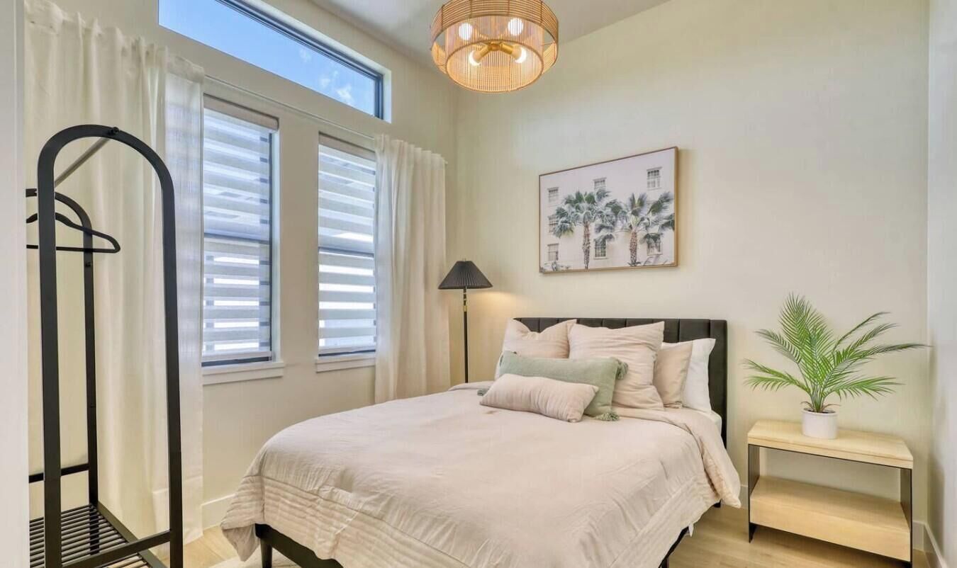 A bedroom with a bed, a bedside table, a window with blinds, a hanging light fixture, and a framed picture on the wall.
