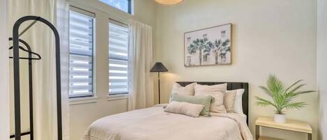 A bedroom with a bed, a bedside table, a window with blinds, a hanging light fixture, and a framed picture on the wall.