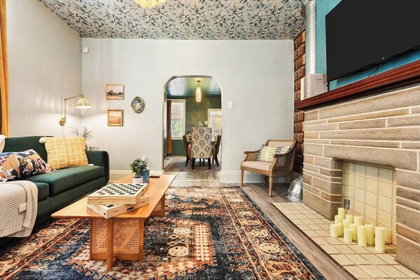 Una sala de estar con un sofá verde, una mesa de centro de madera con un tablero de ajedrez, una alfombra estampada, una chimenea con velas y un área de comedor al fondo.