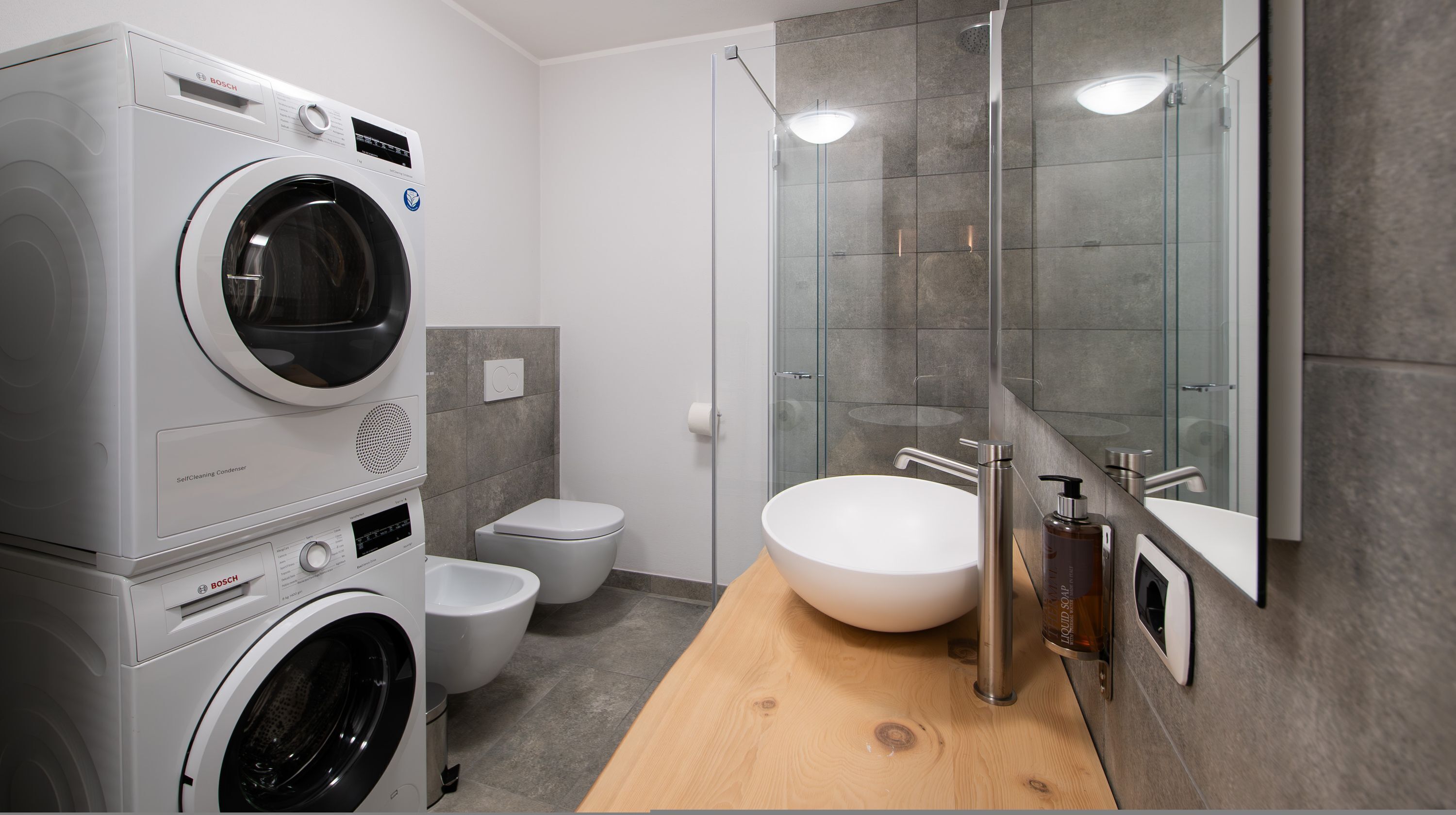 Un baño moderno con una lavadora Bosch, un inodoro, ducha envuelta en vidrio y un tocador de madera con lavamanos.