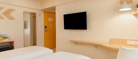 A hotel room with two beds, a TV mounted on the wall, a desk with a chair, and a small shelf with a plant.