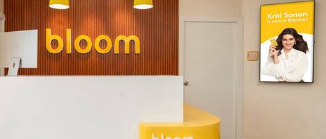 Zona de recepción con pared de madera y un gran cartel con la palabra 'bloom'. Hay un mostrador de recepción amarillo y un cuadro enmarcado en la pared.