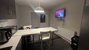 A compact kitchen with a microwave, kettle, and a dining table with chairs. A TV is mounted on the wall, and a ceiling light is visible. - The Coral Guest House (Southampton)