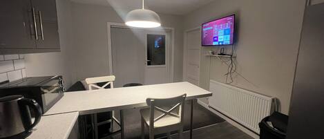 A compact kitchen with a microwave, kettle, and a dining table with chairs. A TV is mounted on the wall, and a ceiling light is visible.