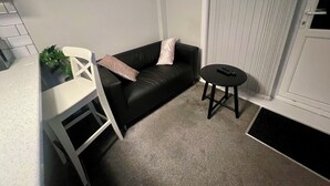 A small, modern living area with a black leather sofa, a white chair, a small round table with a remote control, and a potted plant. - The Coral Guest House (Southampton)