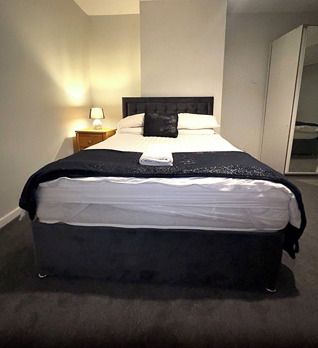 A neatly made bed with a black headboard, a white pillow, and a black pillow on the bed. There is a small bedside table with a lamp and a folded towel on it. The room has a carpeted floor and a door to the right.
