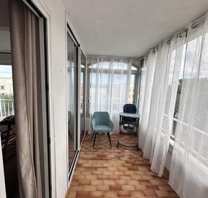 Un balcón con piso de madera, una silla azul, un cochecito de bebé y cortinas de gasa.