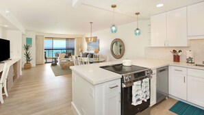 A modern kitchen with white cabinetry, a built-in oven, and a dishwasher. There is a dining area with a table and chairs, and a large window offering a view of the outdoors.