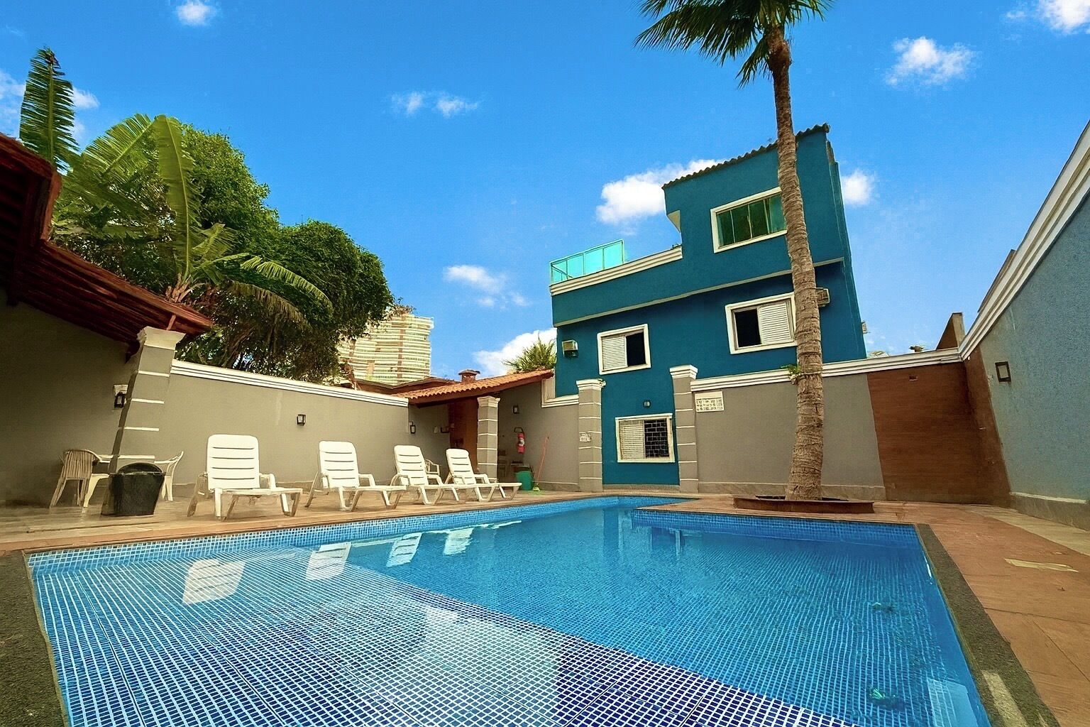 Una piscina con sillones de descanso, una palmera y un edificio azul con ventanas.