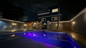 A nighttime swimming pool area with lounge chairs and a building in the background. - Marmeu Enseada com jacuzzi aquecida (Guarujá)