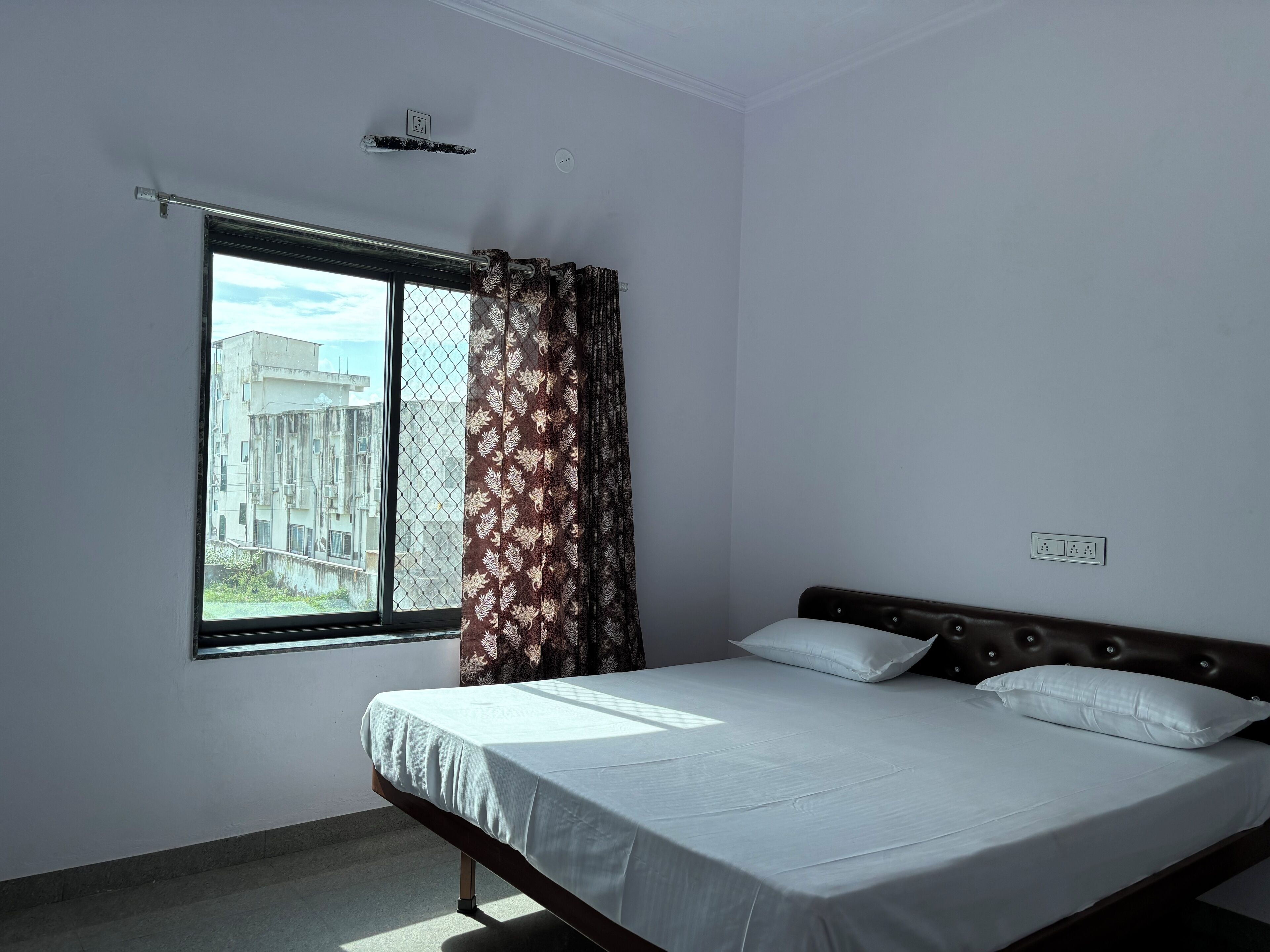 A bedroom with a single bed, a window with patterned curtains, and a view of buildings outside.
