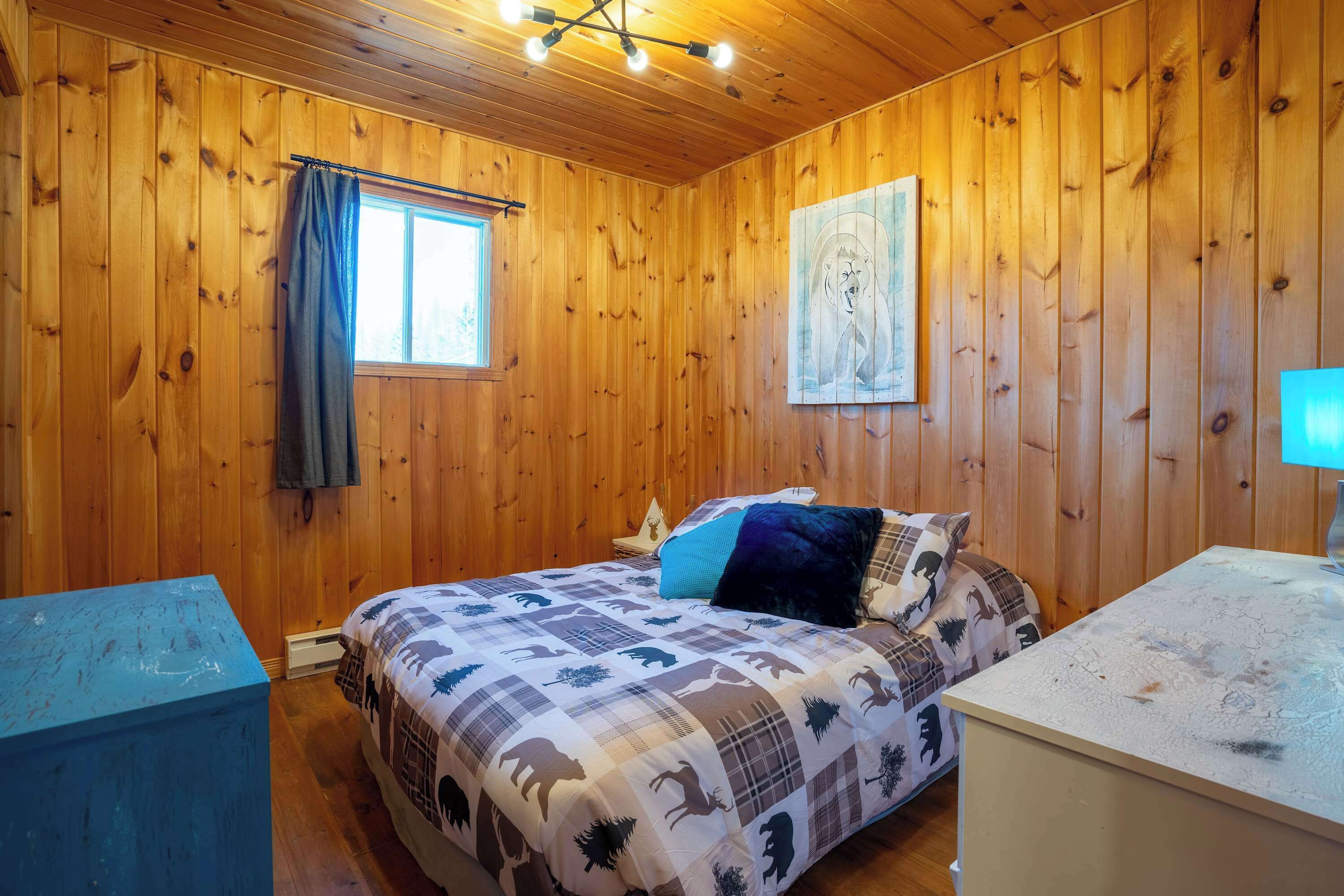 Ein Zimmer mit Holzwänden, einem Bett, einem Nachttisch, einem Fenster mit Vorhang und einem Bild an der Wand.