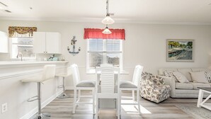 Una cocina luminosa con armarios blancos, un comedor con sillas blancas y una sala de estar con sofá y cojines.