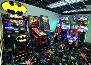Une salle avec des jeux d’arcade, notamment des jeux sur le thème de Batman et des jeux de course, et un sol décoré de motifs en forme d’étoiles.